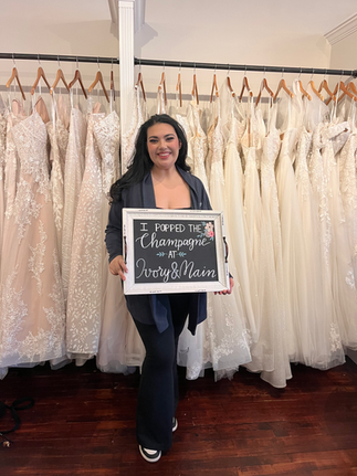 a girl stands in a bridal shop holding a sign that says "I popped the champagne at Ivory & Main"