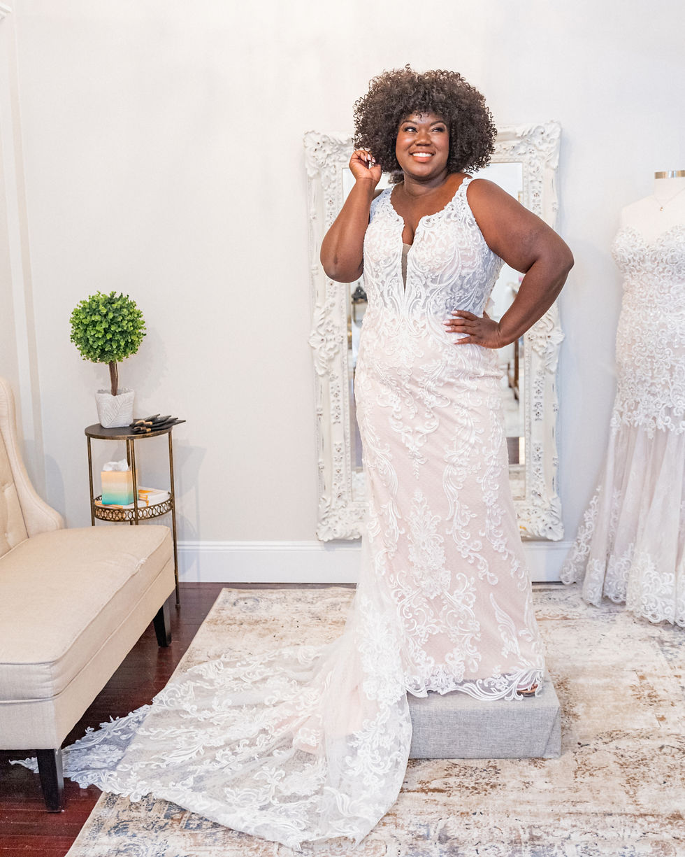 Woman smiling in an ornate white lace wedding dress, standing on a pedestal in a bright room. Beige chair and greenery in background.