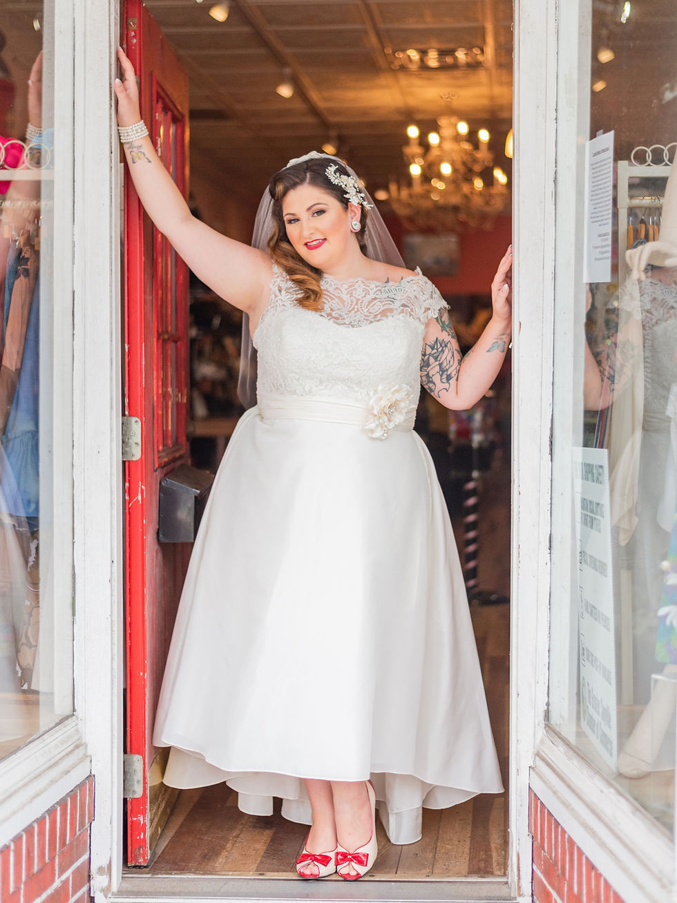 Bride in white dress with lace top and red shoes stands in doorway, smiling. Indoor chandelier lights and red door create a warm setting.