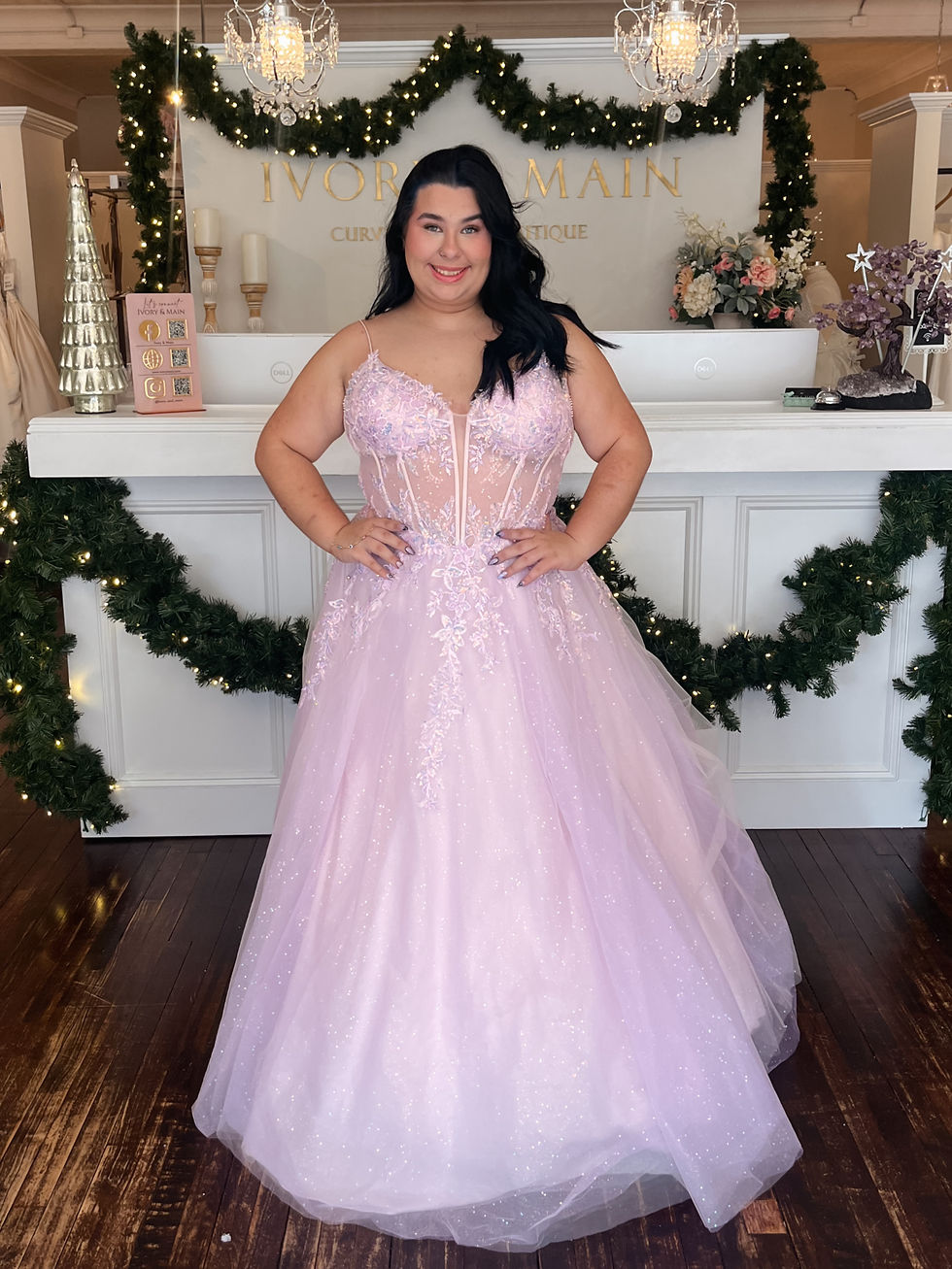 Woman in a pink prom gown smiles at a decorated boutique. Background has garlands and chandeliers. Sign reads "Ivory Main Curvy Boutique."