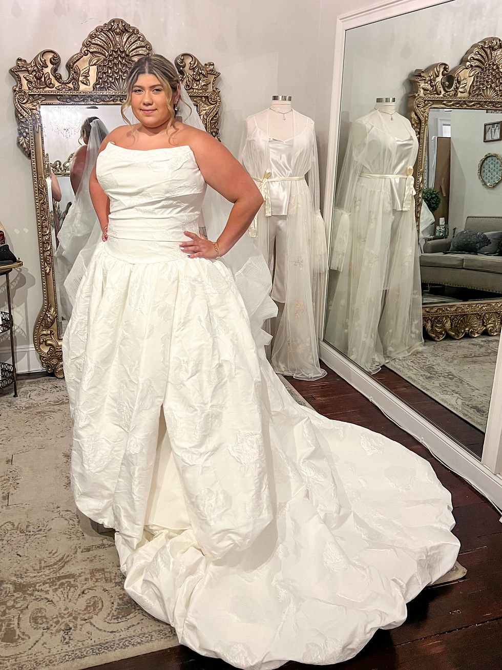 Bride in a strapless white wedding gown poses in a bridal boutique, with ornate mirrors and mannequin dresses behind her.