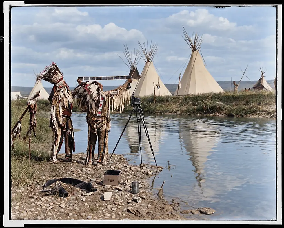 Two Native men in feathered regalia examine photographic film beside a camera and tripod near a stream, with tipis in the background.