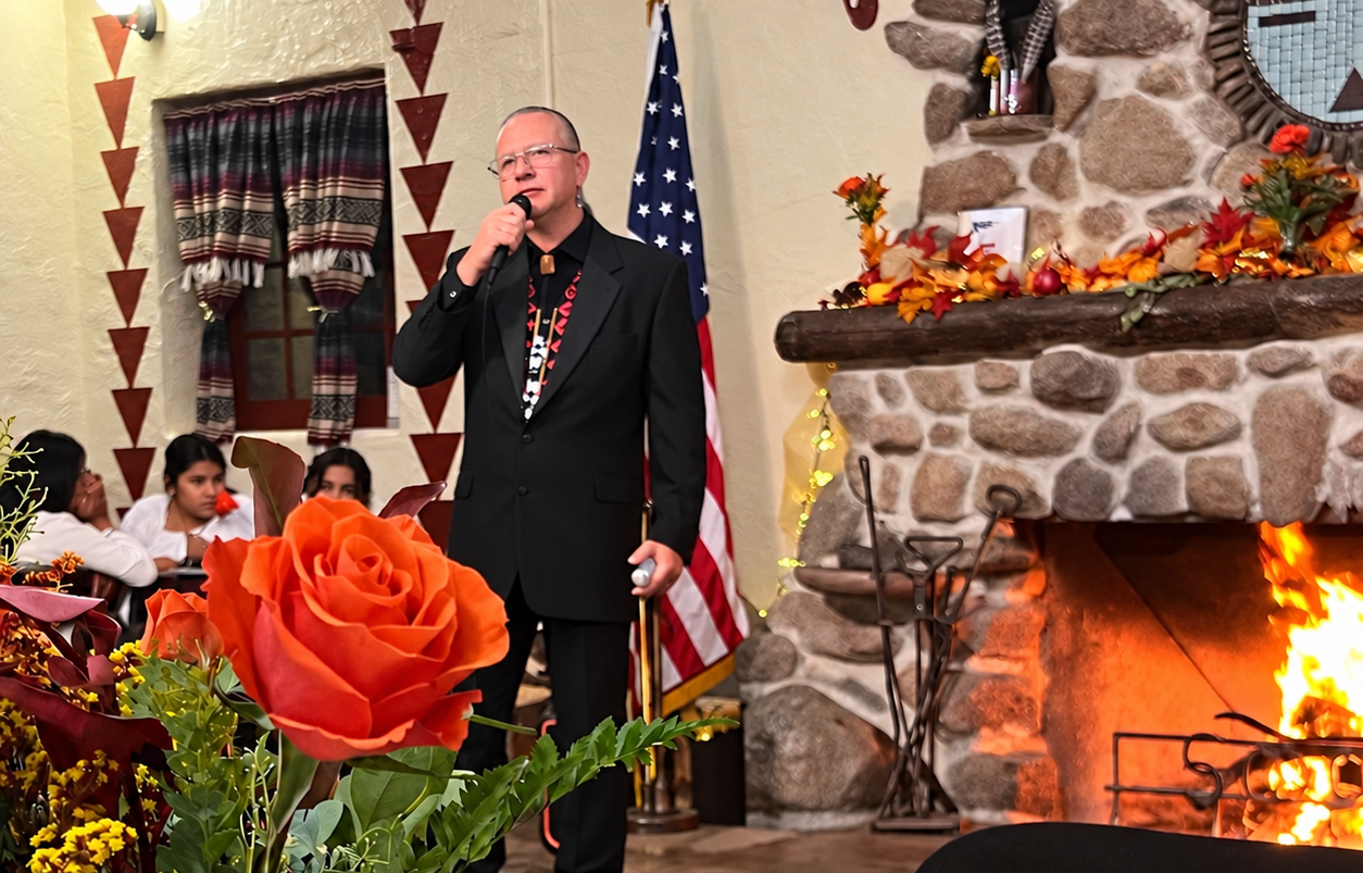 MIP Executive Director Manuel Lucero addresses guests during the 2025 First Fire Fall Fundraiser inside the museum's historic Pueblo Building. Autumn floral arrangements, a decorated stone fireplace, and tables prepared for dinner create a welcoming setting for the annual celebration.