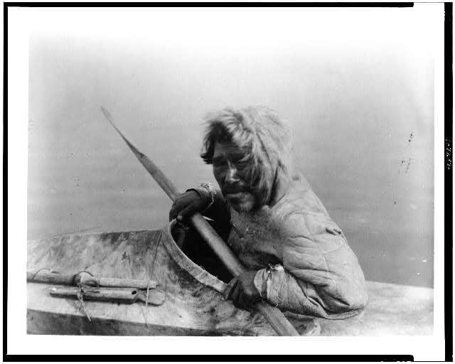 Black-and-white photograph of a Noatak seal hunter seated in a kayak while holding a paddle.