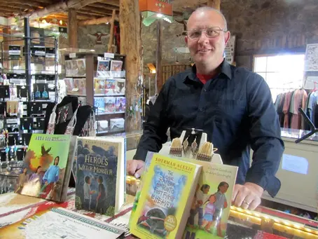 Manuel Lucero stands behind the trading post counter at the Museum of Indigenous People, surrounded by Native-authored books, jewelry, and handmade Indigenous art.
