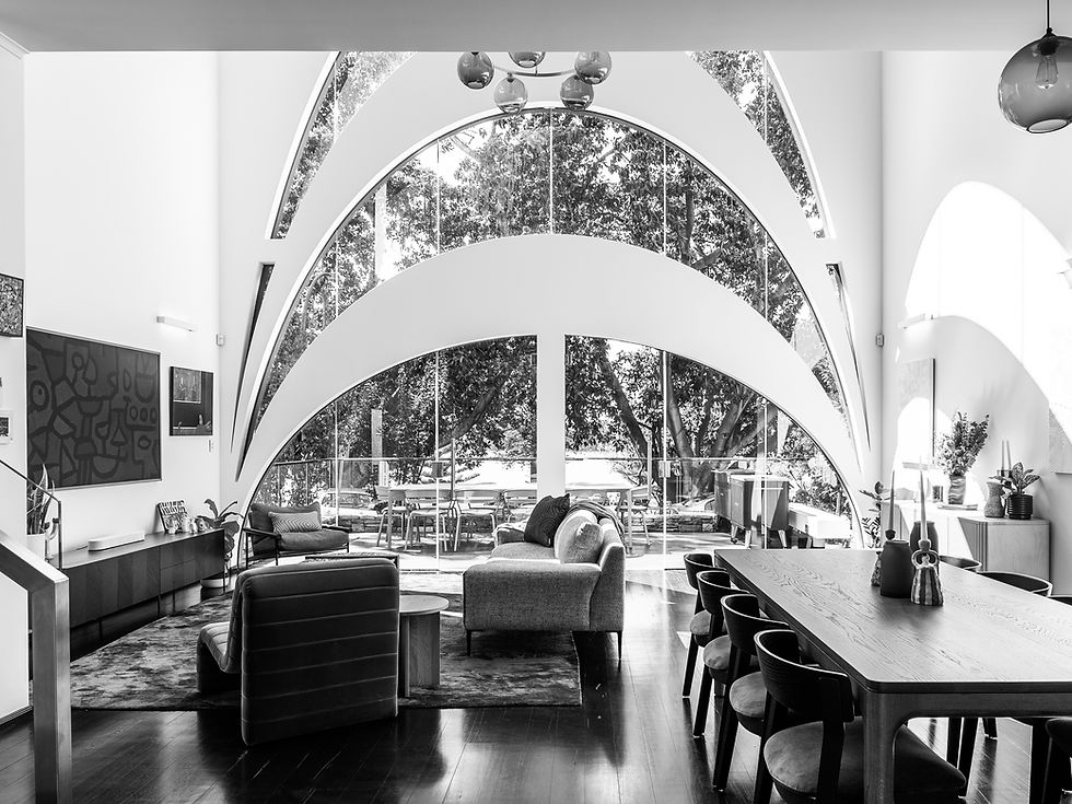 Black and white interior photo of a modern living room with arched ceiling and large arched windows. The room has contemporary furniture and artwork, with trees visible through the windows.
