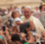 Pope Francis, surrounded by a crowd, smiles as a young child is lifted toward him.