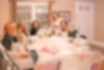 Five women smiling at a table, each holding decorated cookies. The setting is cozy with a white tablecloth and wrapped gifts, creating a festive mood. Sarah Grace Roberts Sarah Grace Cookie Co.