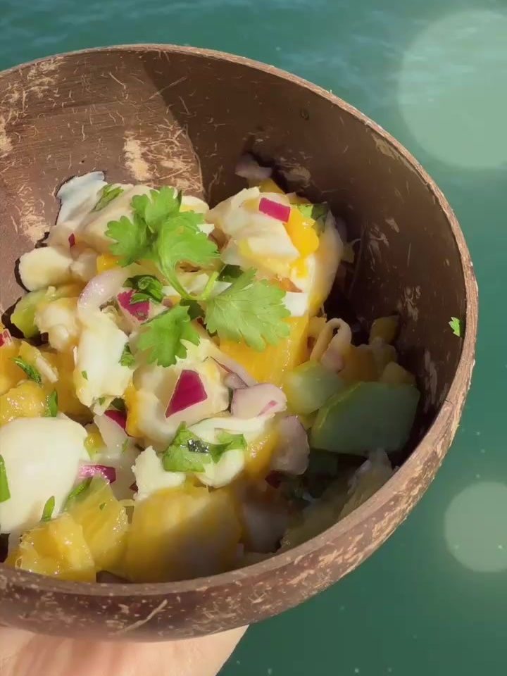 Mango, coconut, and pineapple ceviche with cilantro and red onion, served in a coconut bowl over turquoise water.