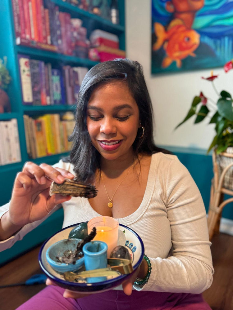 dominican artist and coach, Jasleni, looking down at her hands, holding sage, bookshelf behind her