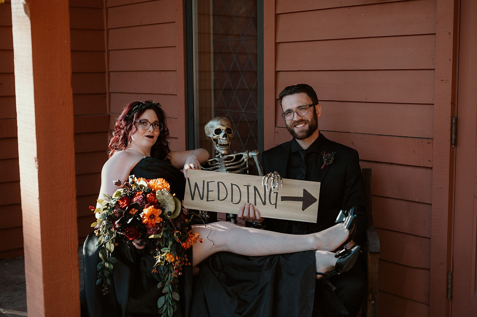 Couple with skeleton sits on a bench in black attire, holding a "Wedding" sign. The woman holds a vibrant bouquet. Mood is playful.