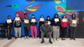 A group of people hold certificates, smiling in front of a vibrant mural featuring a colorful hummingbird and floral patterns on a blue wall.