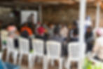 A group of women sits in a circle on white chairs indoors, engaged in discussion. One gestures as others listen attentively. Brick walls surround them.