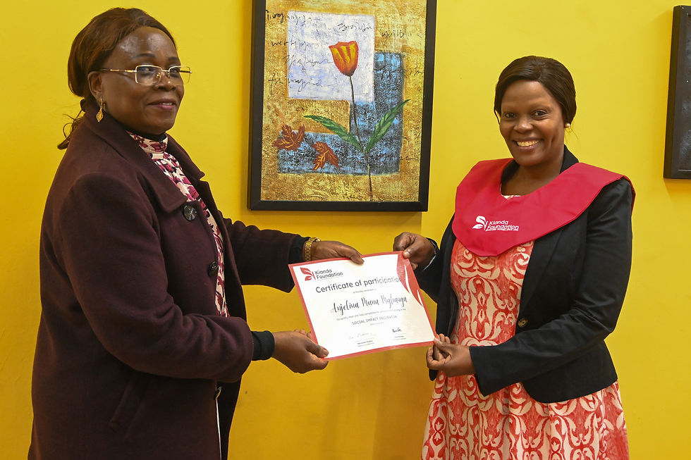 Two women stand smiling. One hands a certificate to the other. They're in front of a bright yellow wall with artwork.