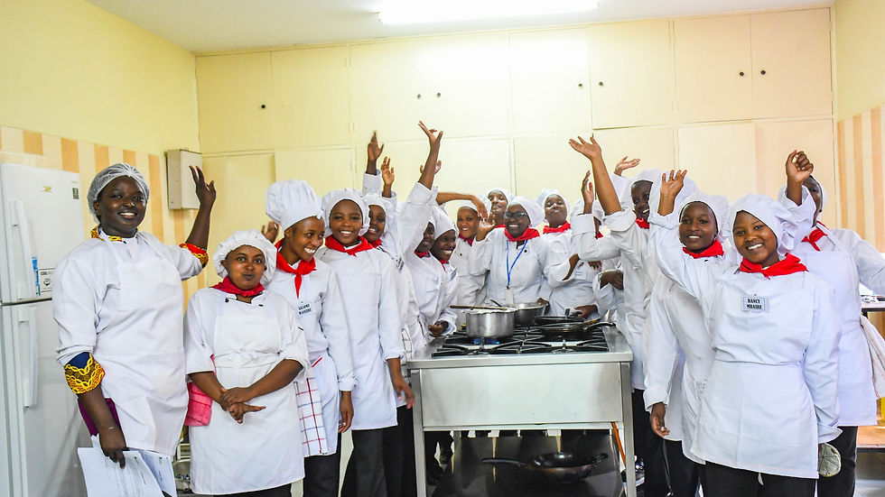 Chefs in white uniforms and red scarves smile and raise hands in a bright kitchen, standing around a stove; a joyful, celebratory mood.