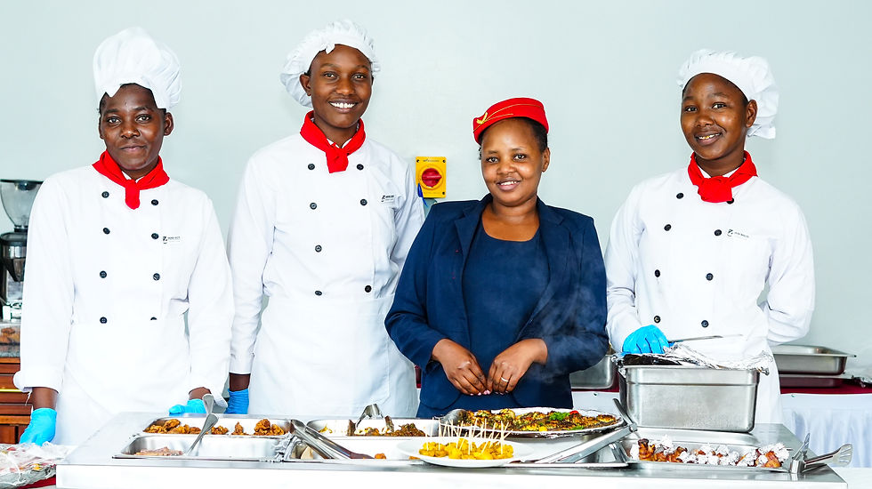 Chefs in white uniforms with red scarves stand smiling beside a woman in a navy blazer. They are in a kitchen with food trays.