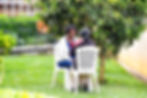 Two women sit on white chairs in a garden, engaged in conversation under a tree. The scene is calm, with greenery and a railing nearby.