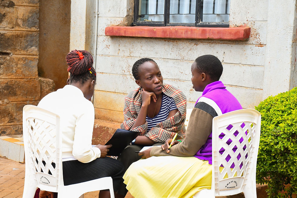 Three people sit on white chairs outside a brick building, engaged in discussion. One holds a notebook. Bright clothing and greenery nearby.