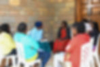 A group of women sit on white chairs in discussion near a brick wall. One takes notes, others listen attentively. Casual and engaged mood.