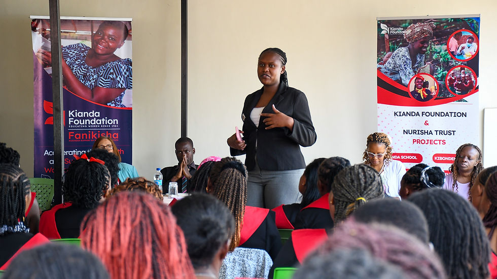 A woman speaks to an audience with black and red attire. Two banners promote Kianda Foundation and Nurisha Trust. Bright, engaging atmosphere.