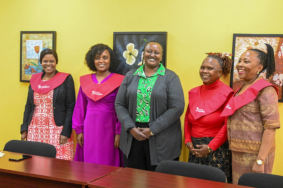 Five smiling women in colorful attire and red sashes stand in a room with yellow walls and framed art. A table with a phone is in front.
