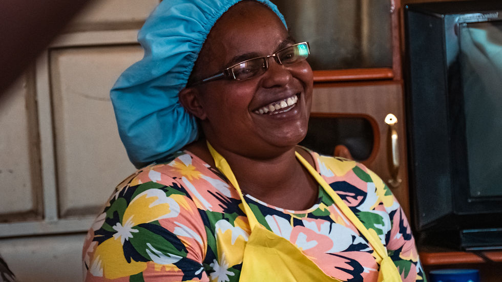 Smiling woman in floral shirt and yellow apron, wearing a blue bonnet, sits in a cozy room. Warm atmosphere with a TV in the background.
