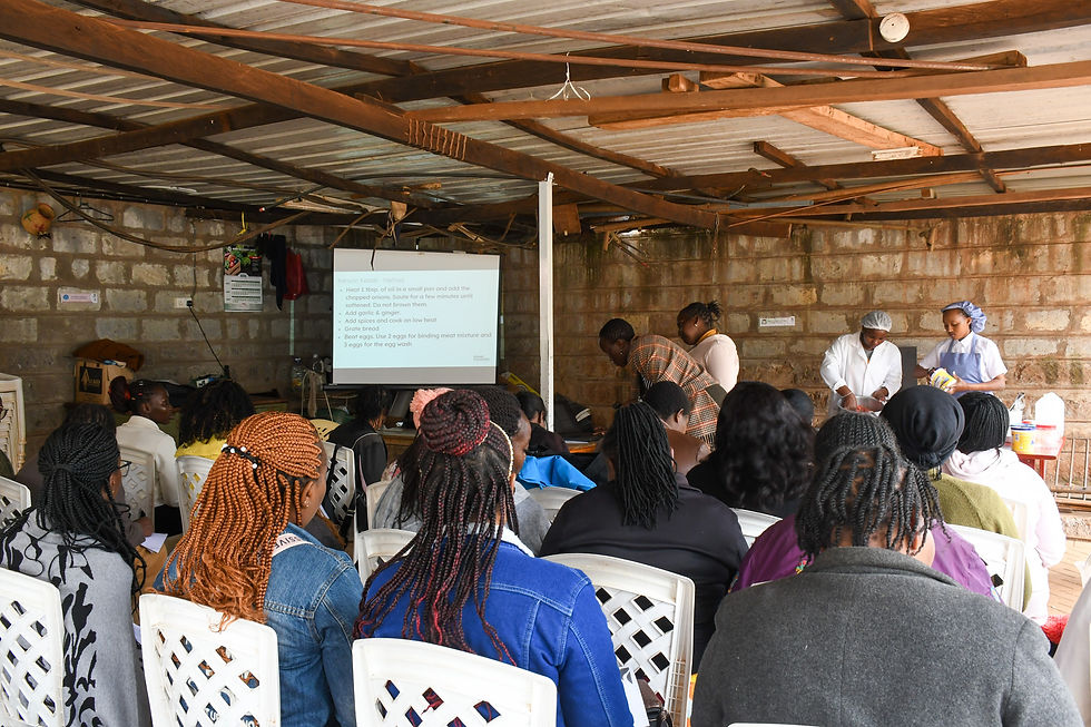 Audience seated in a rustic room watching a cooking presentation. A screen displays a kebab recipe. Instructors prepare ingredients.