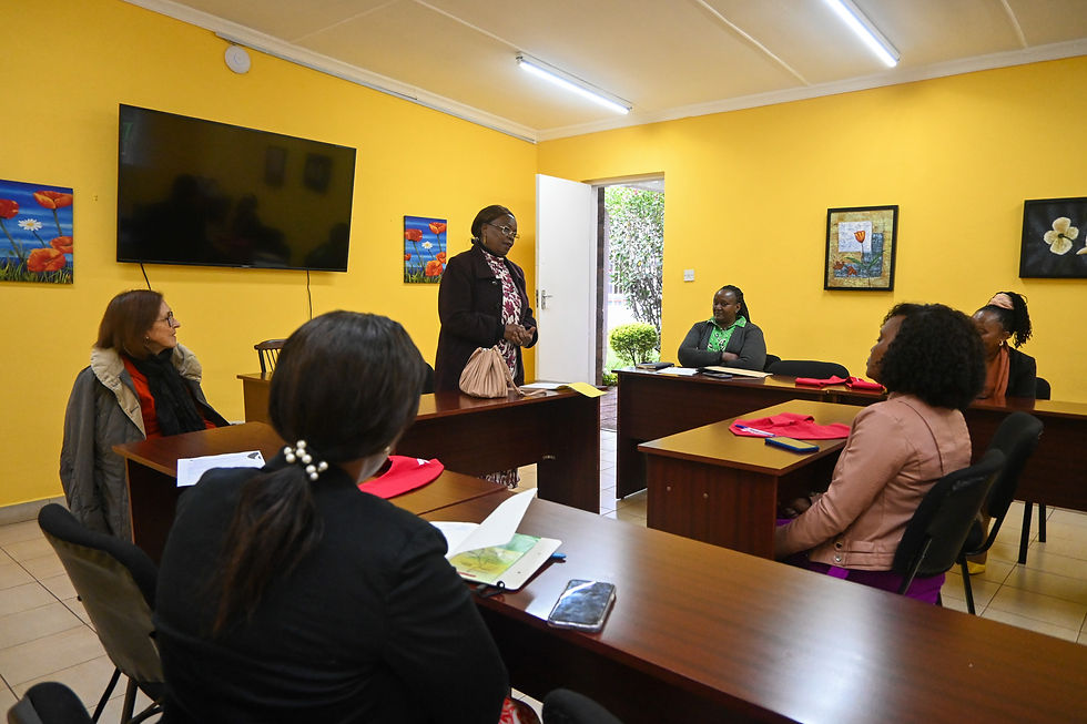 A woman stands speaking to five seated women in a bright yellow room with floral paintings and a TV. The setting is a classroom.