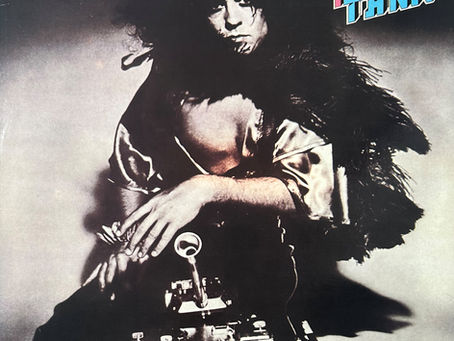 Marc Bolan crouches over a toy tank in a dramatic black‑and‑white photograph, feather boa draped over his shoulders, the tank’s barrel pointed toward the viewer — a surreal, theatrical image that defines the Tanx era.
