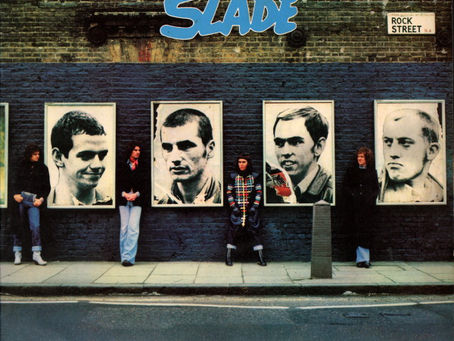 Album cover showing Slade members standing beneath five large black‑and‑white portrait posters on a brick wall, with the title Whatever Happened to Slade above in stylised lettering and a street sign reading “ROCK STREET N4.”