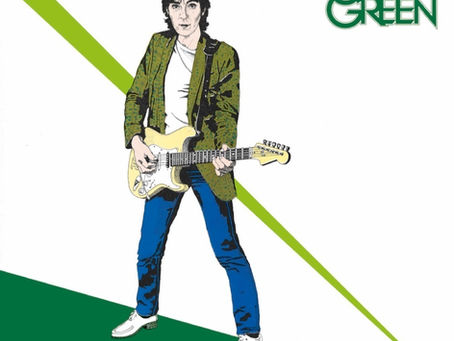 A black‑and‑white photograph of Jack Green holding a guitar, mid‑1970s, wearing a patterned shirt and standing in a rehearsal space with amplifiers behind him.