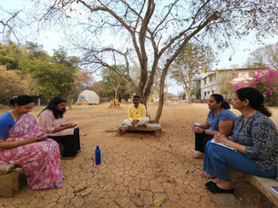 Five people sit in a circle outdoors under a large tree, taking notes in a dry park with buildings and pink flowers.