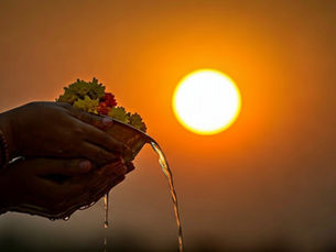 Hands holding a bowl of flowers pour water, silhouetted against a vivid orange sunset. The scene conveys serenity and reflection.