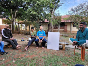 Group seated outdoors in a yard, chatting around a blank whiteboard; man in blue Dream11 India jersey and relaxed smiles.