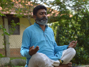 Bearded man in blue kurta meditates cross-legged outdoors with eyes closed, in a leafy garden by a house.