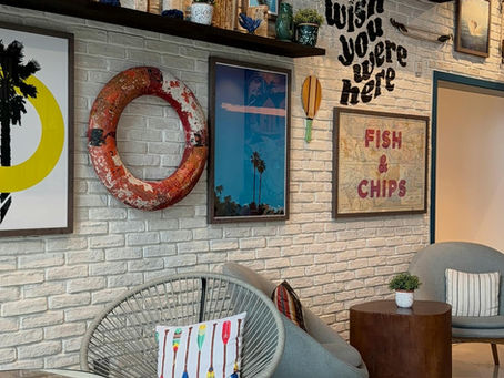 Coastal-inspired vacation rental living room with a curated selfie wall featuring beach decor, framed artwork, floating shelves, and a cozy sectional sofa designed to create a memorable guest experience and boost direct booking appeal.