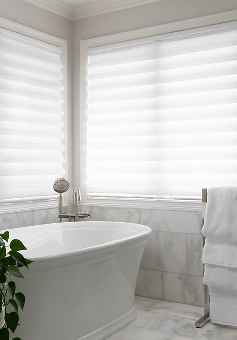Serene primary bathroom with a freestanding soaking tub, sheer roman shades, and marble tile.
