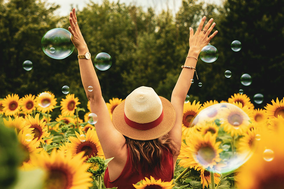 A person in a hat raises arms in a sunflower field with bubbles floating, under a bright sky, evoking joy and freedom.