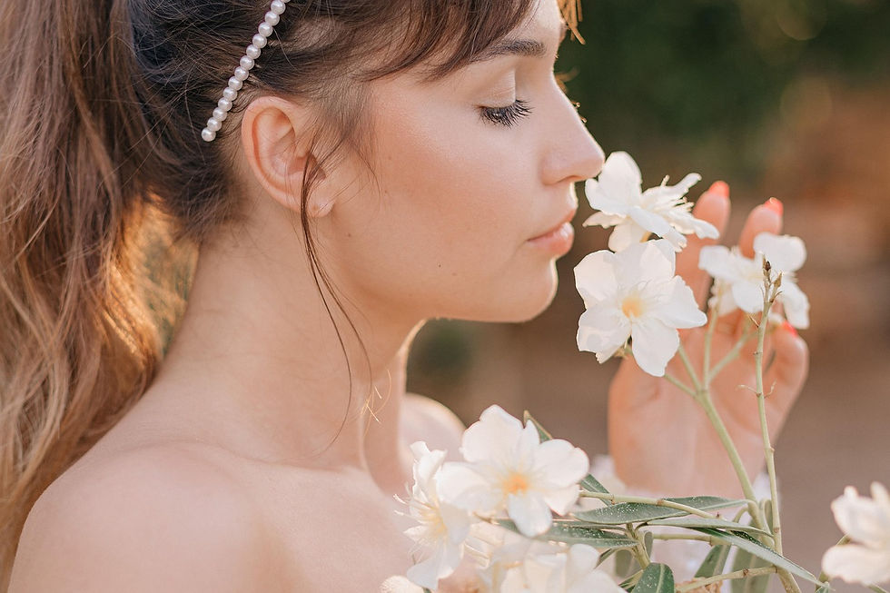 Woman with pearl hairpin gently smells white flowers in sunlight, conveying tranquility. Background is blurred with soft, warm tones.