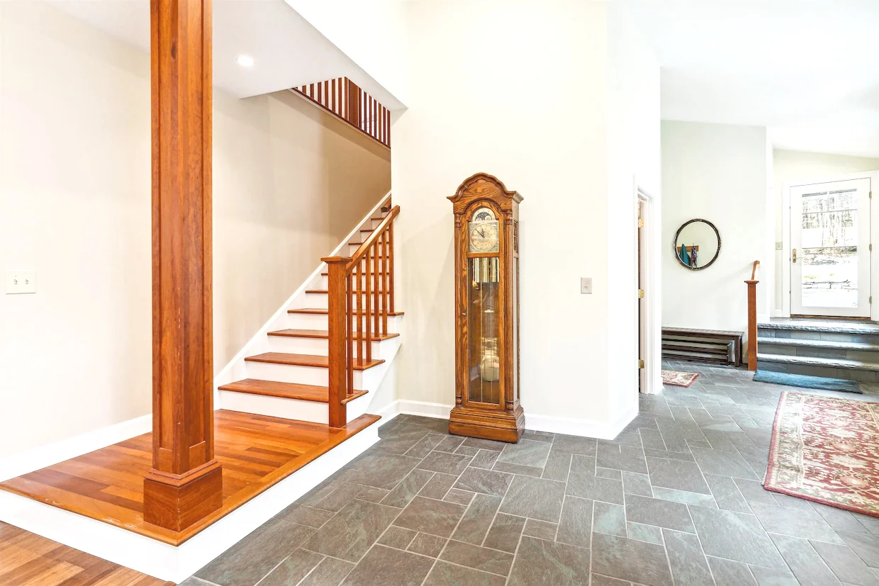A tiled entry hall with a wood stair, tall grandfather clock, and doorways into adjoining rooms.