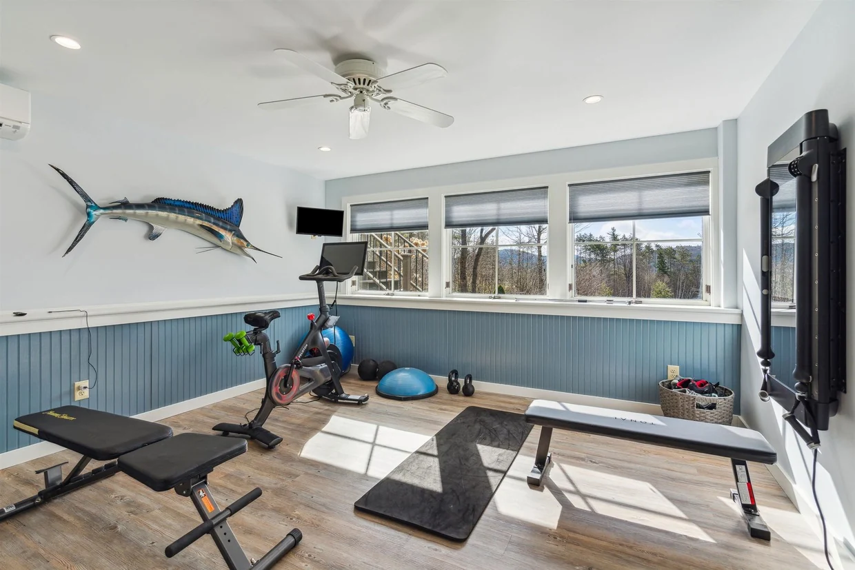 A finished exercise room with windows, a weight bench, fitness equipment, and blue lower wall paneling.