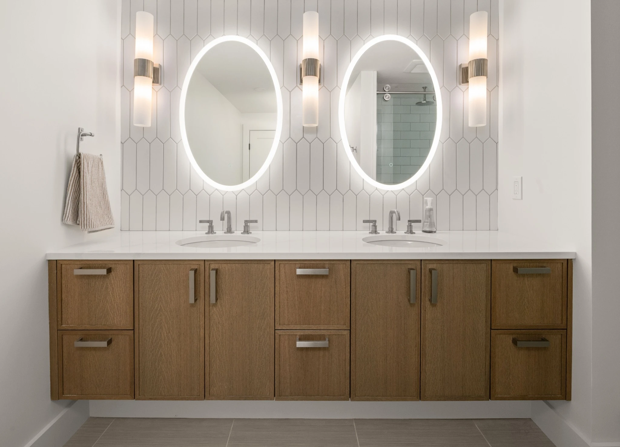 Illuminated oval mirrors and wall sconces above a wood vanity and geometric tile.