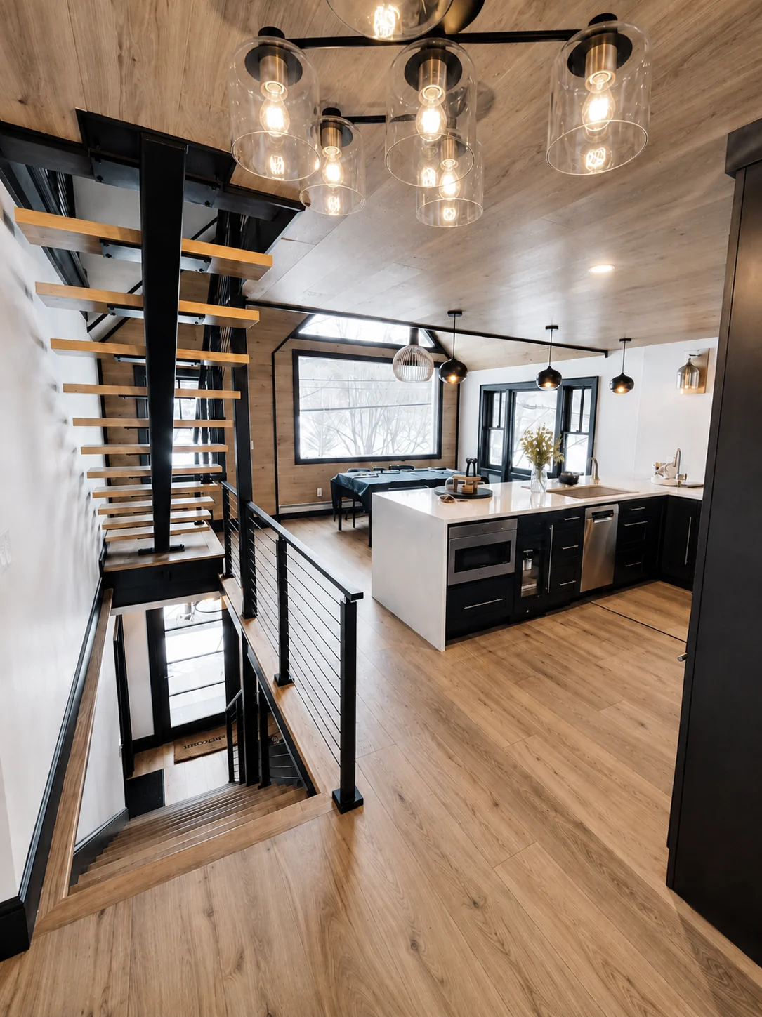 A tall view through the kitchen beside an open wood staircase, with pendant lights and a dining table beyond.