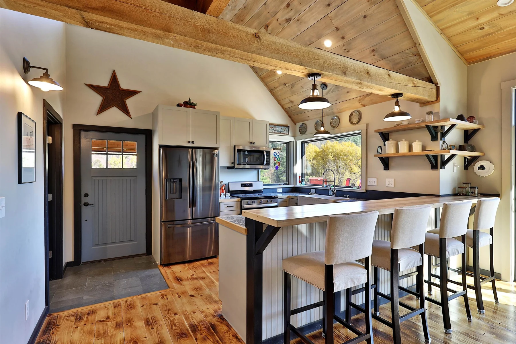 A pale kitchen island with stools beneath exposed timber framing, beside the refrigerator and wood shelving.
