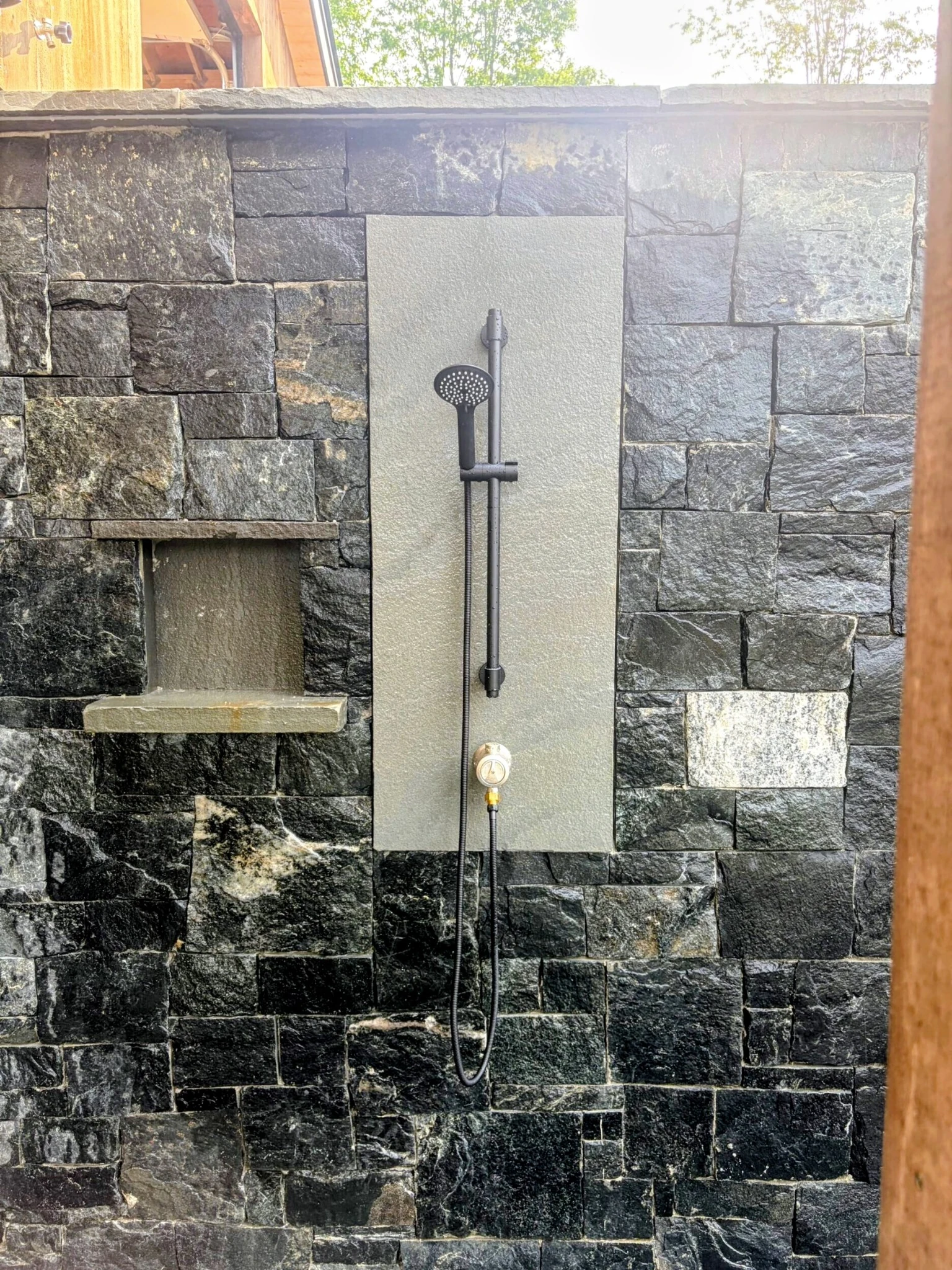 A dark handheld outdoor shower against natural stone, with an inset shelf and timber framing at the Rochester home.