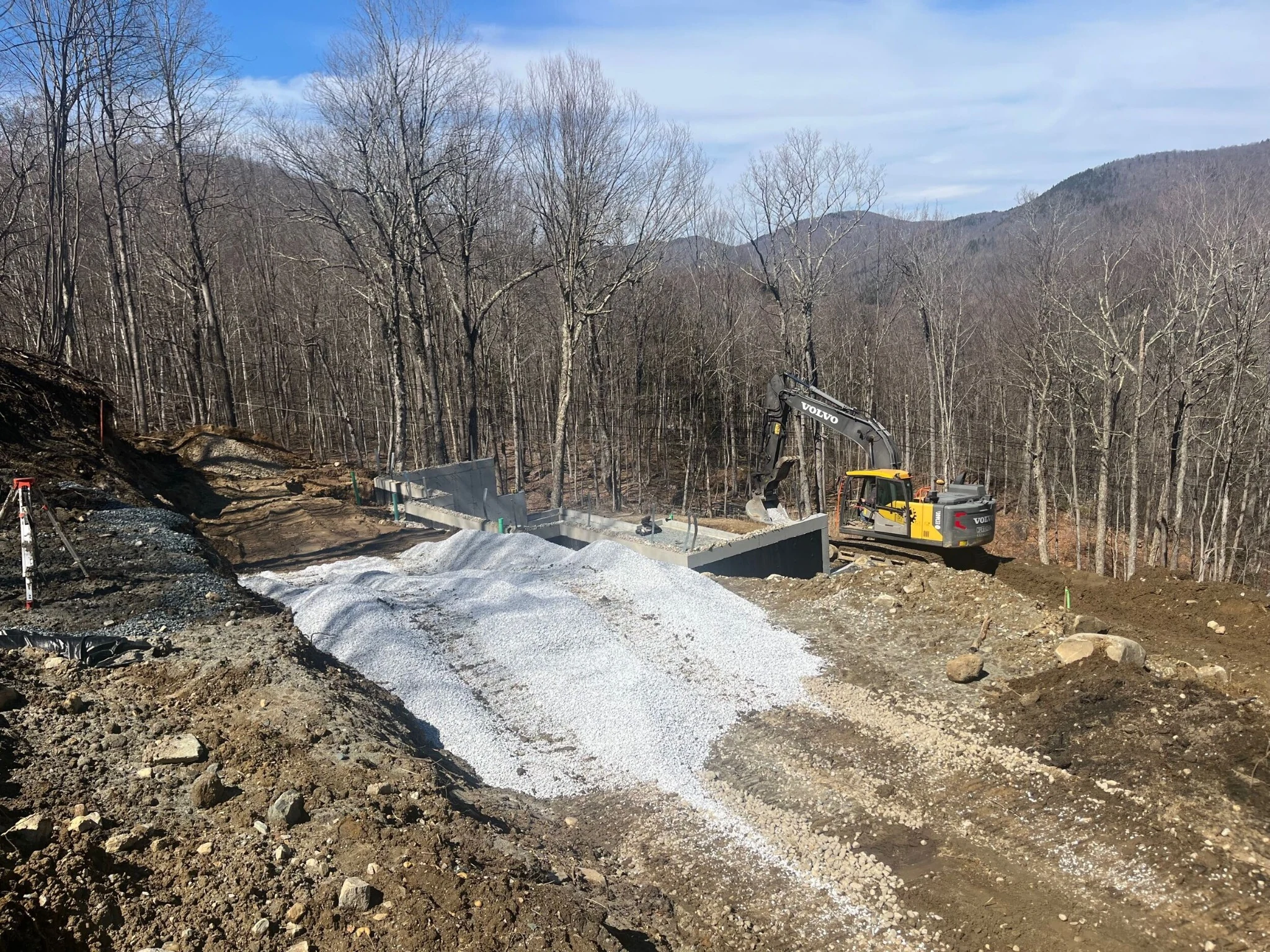 Excavation and concrete foundation walls on a sloping wooded site, with gravel and an excavator.