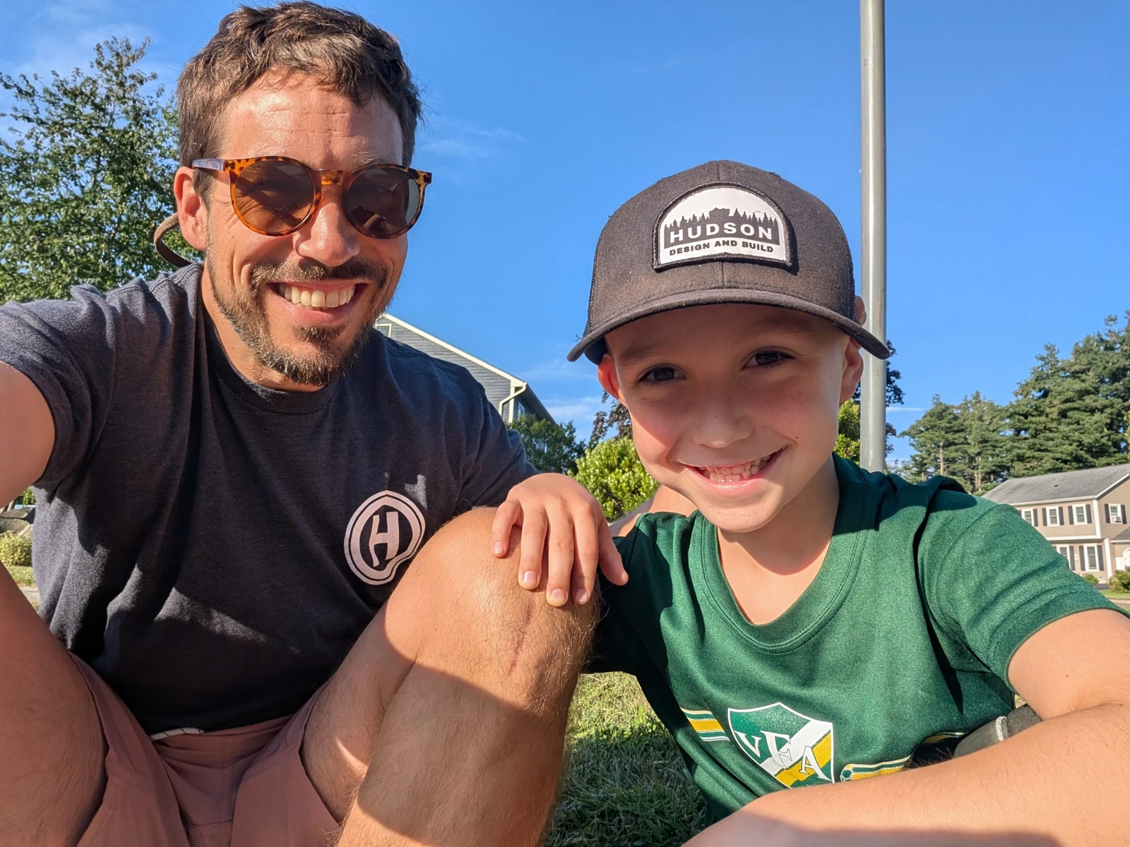 An adult and child smiling outdoors, with Hudson branding on a shirt and cap.
