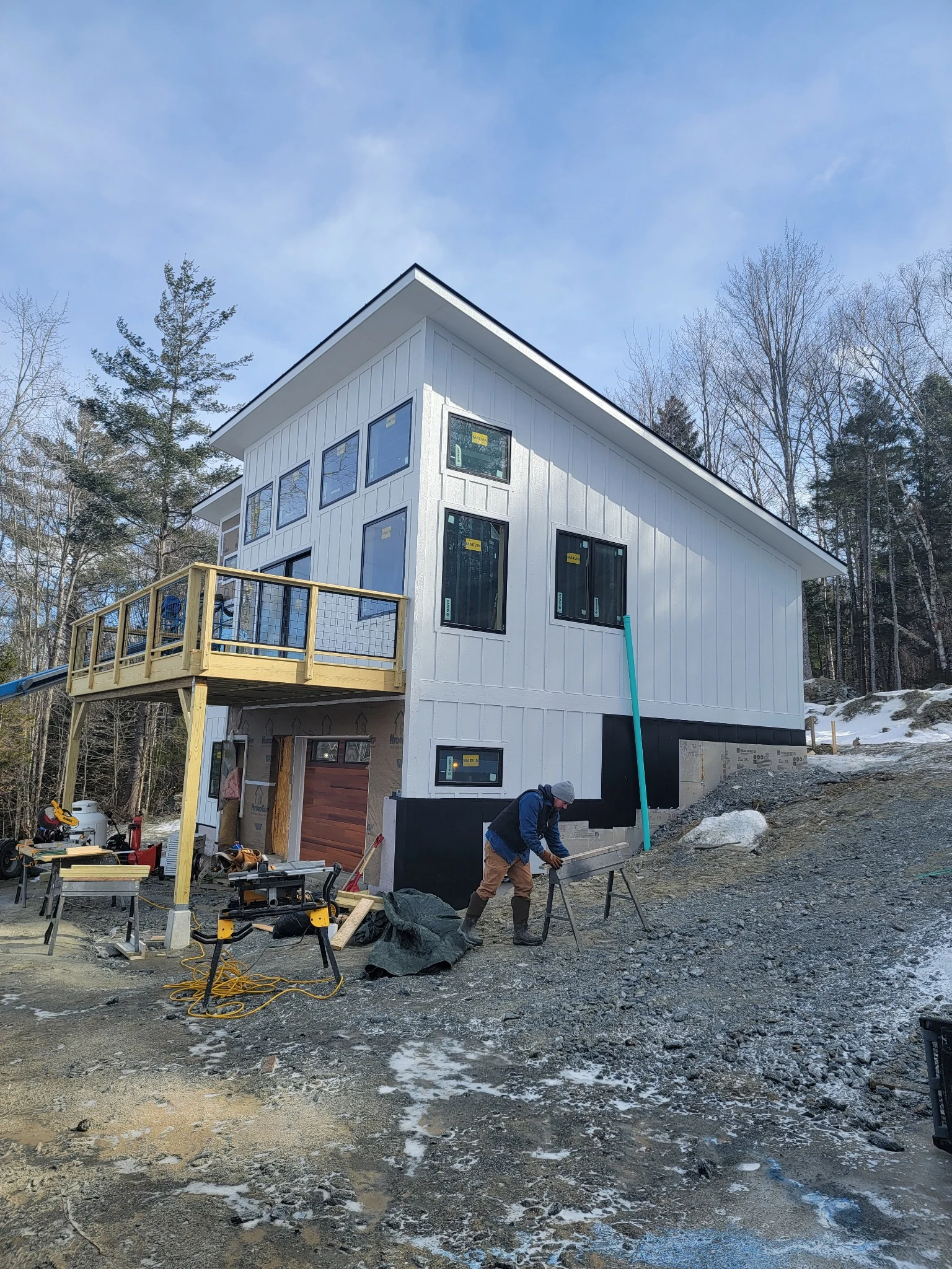 A Hudson carpenter working outside a white home under construction, with tools, a new deck and snow on the wooded slope.