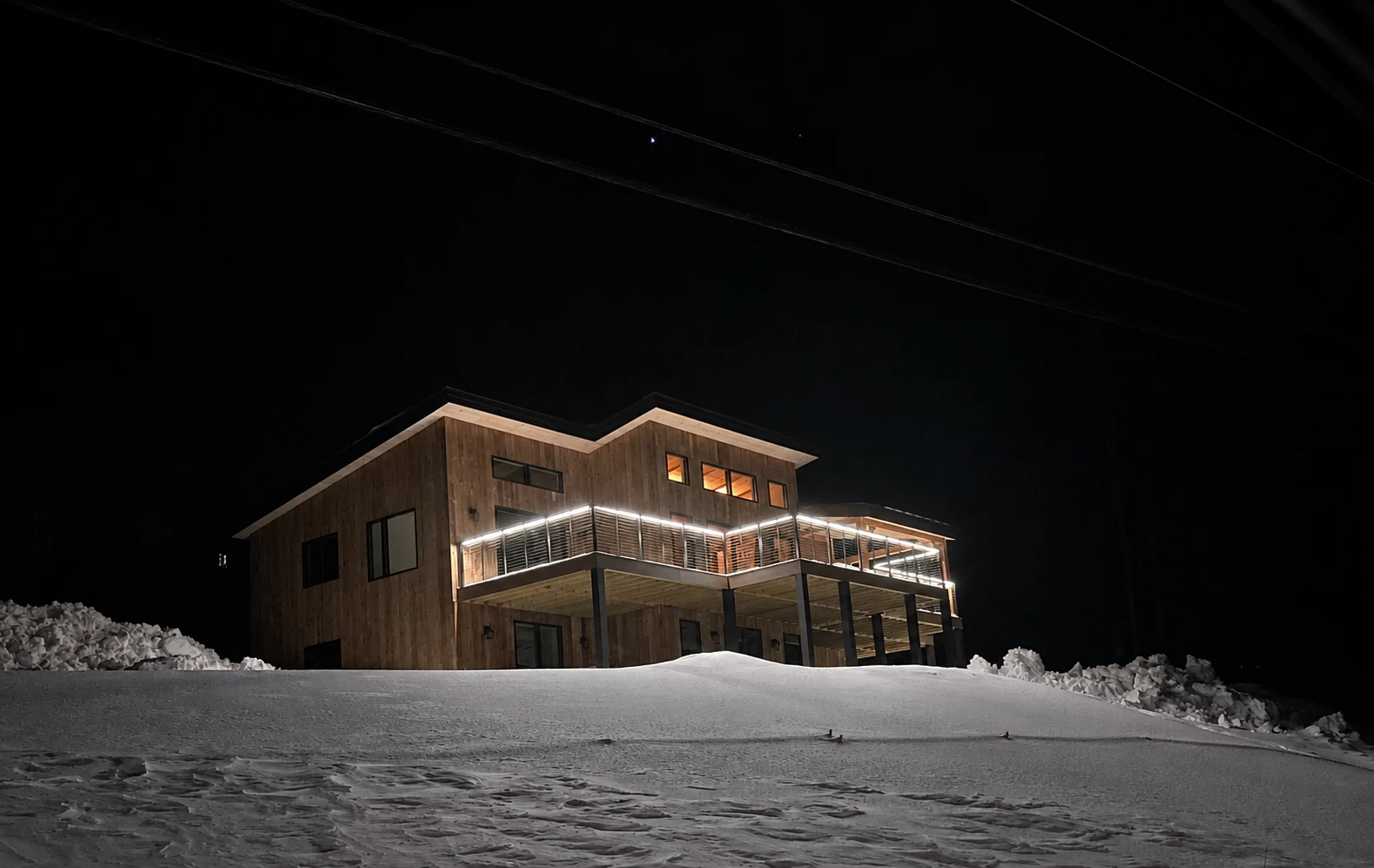 Linear deck railing lights and warm windows on a wood-clad mountain home above snow at night.