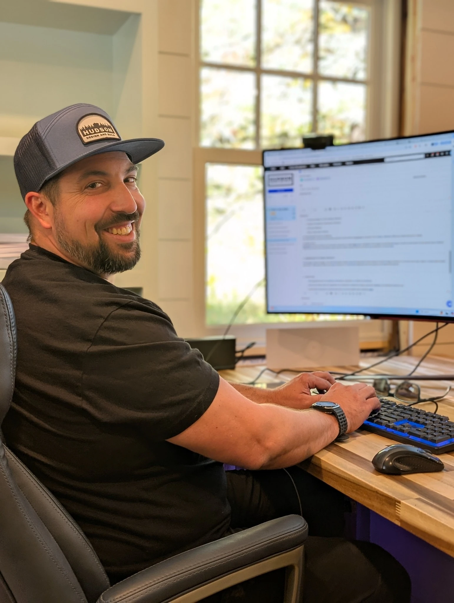 A smiling Hudson team member at his desk, wearing a Hudson cap.